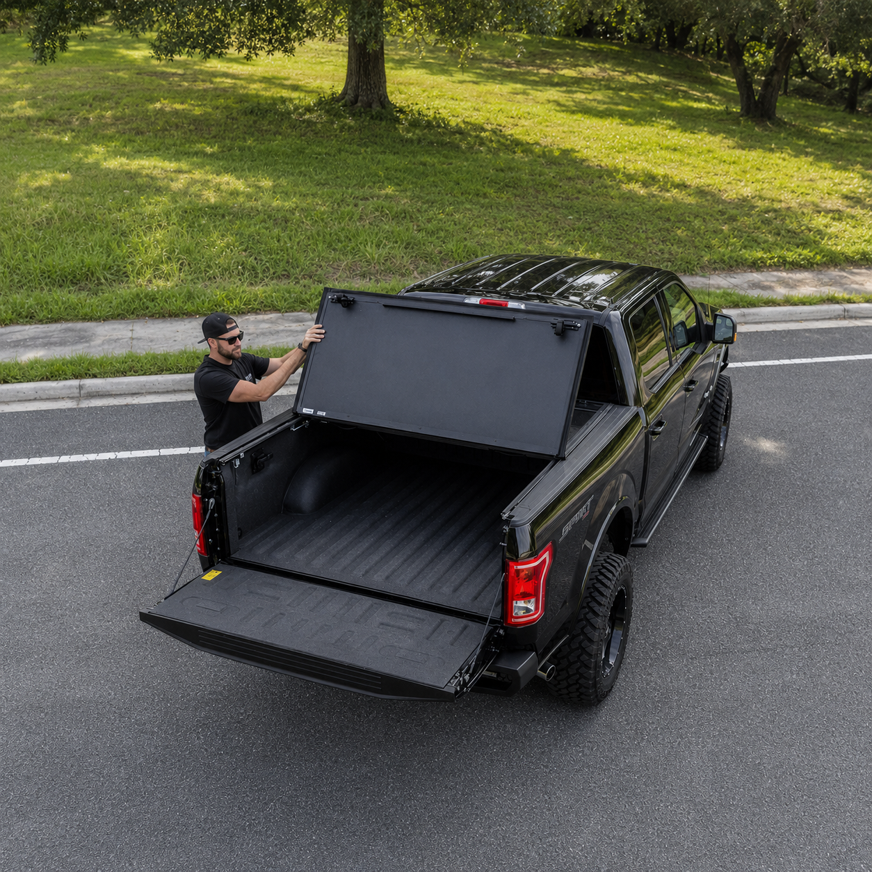 Man opening the tailgate of a black truck on a street with grass and trees in the background